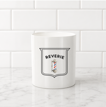 Load image into Gallery viewer, A white ceramic mug with a black-outlined shield graphic reading 'REVERIE' above a barber pole illustration, set on a white marble surface against a white subway tile background.