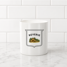 Load image into Gallery viewer, A white ceramic mug with a black-outlined shield graphic featuring the word 'REVERIE' above a small illustrated cabin scene, set on a marble surface against a white subway tile background.
