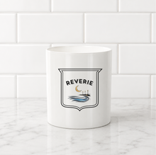 Load image into Gallery viewer, A white ceramic mug with a black and blue 'Reverie' shield graphic, set on a white marble countertop against a white subway tile backsplash.