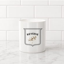 Load image into Gallery viewer, A white ceramic mug with a black and gold shield logo reading 'REVERIE' sits on a white marble countertop against a white subway tile backsplash.