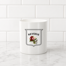Load image into Gallery viewer, A white ceramic mug with a black shield graphic reading 'REVERIE' above a small floral illustration, set on a white marble surface against a white subway tile background.