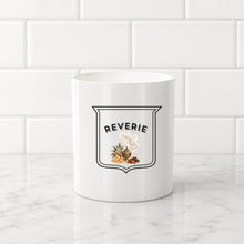 Load image into Gallery viewer, A white ceramic mug with a black and white shield graphic reading 'REVERIE' and a small illustrated cluster of herbs, set on a white marble countertop against a white subway tile backsplash.