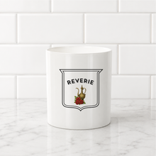 Load image into Gallery viewer, A white ceramic mug with a black and red 'Reverie' shield graphic, set on a white marble countertop against a white subway tile backsplash.