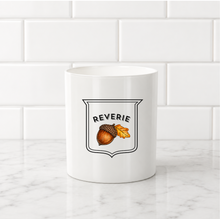 Load image into Gallery viewer, A white ceramic mug with a 'REVERIE' graphic featuring an acorn and autumn leaves, set on a white marble countertop against a white subway tile backsplash.
