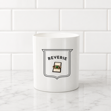 Load image into Gallery viewer, A white ceramic mug with a black shield graphic reading 'REVERIE' and a small green-and-brown illustration, set on a white marble surface against a white subway tile wall.
