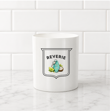 Load image into Gallery viewer, A white ceramic mug with a 'Reverie' cocktail graphic, set on a white marble countertop against a white subway tile backsplash.
