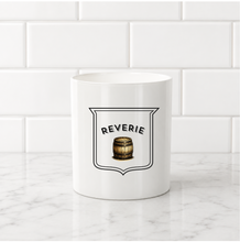 Load image into Gallery viewer, A white ceramic mug with a black and brown 'Reverie' barrel graphic, set on a white marble countertop against a white subway tile backsplash.
