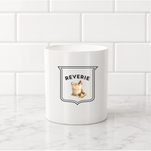 Load image into Gallery viewer, A white ceramic mug with a black and gold 'Reverie' cocktail graphic, set on a white marble countertop against a white subway tile backsplash.
