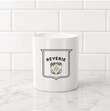 Load image into Gallery viewer, A white ceramic mug with a black and gold shield emblem featuring the word 'REVERIE' and a floral crest, set on a white marble countertop against a white subway tile backsplash.
