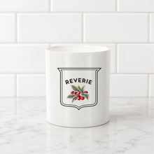 Load image into Gallery viewer, A white ceramic mug with a vintage-style 'Reverie' shield emblem featuring red berries and green leaves, set on a white marble countertop against a white subway tile backsplash.
