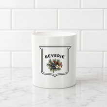 Load image into Gallery viewer, A white ceramic mug with a black-outlined crest and a Christmas wreath graphic featuring the word 'REVERIE' in arched black text, set on a white marble countertop against a white subway tile backsplash.
