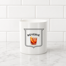 Load image into Gallery viewer, A white ceramic mug with a black-outlined shield graphic reading 'REVERIE' above an illustration of an amber cocktail, set on a marble surface against a white subway tile background.
