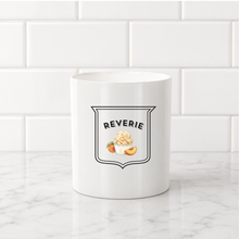 Load image into Gallery viewer, A white ceramic mug with a black and white shield logo that reads 'REVERIE' above a small peach and cream graphic, set on a white marble countertop against a white subway tile backsplash.

