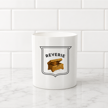 Load image into Gallery viewer, A white ceramic mug with a black and gold treasure chest graphic and the word 'REVERIE' printed on the front, set on a white marble countertop against a white subway tile backsplash.
