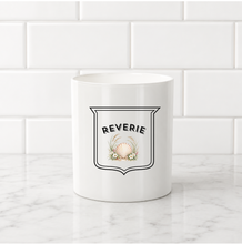 Load image into Gallery viewer, A white ceramic mug with a black outlined shield logo reading 'REVERIE' and a small shell illustration, set on a white marble surface against a white subway tile background.

